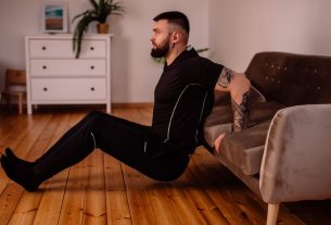 A bearded man in black fitness attire exercising at home, showcasing arm tattoos and a healthy lifestyle.